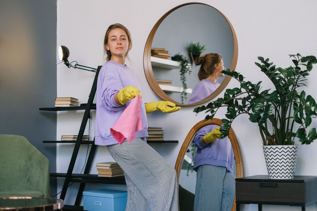 Side profile of a cheerful woman in casual clothes and latex gloves standing beside mirrors while cleaning an apartment during the day